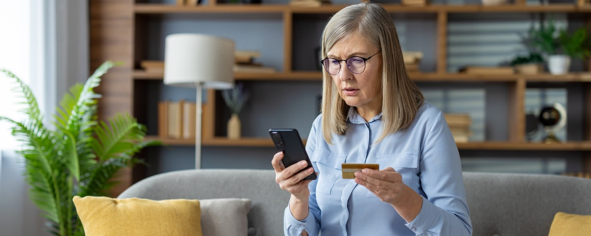 Femme d'âge mûr tenant une carte de crédit et un téléphone, regardant l'écran avec inquiétude.